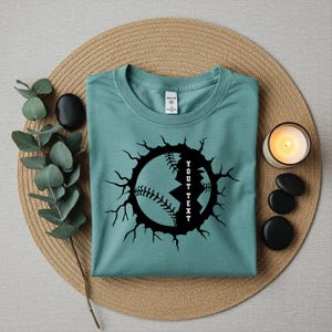 May include: A teal t-shirt featuring a black baseball graphic with a cracked effect. The text "YOUT TEXT" is vertically inside the baseball design. The shirt is folded on a woven mat, with a candle and decorative stones.