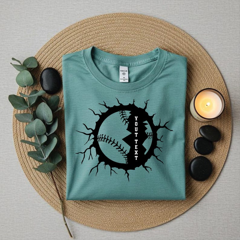 May include: A teal t-shirt featuring a black baseball graphic with a cracked effect. The text "YOUT TEXT" is vertically inside the baseball design. The shirt is folded on a woven mat, with a candle and decorative stones.
