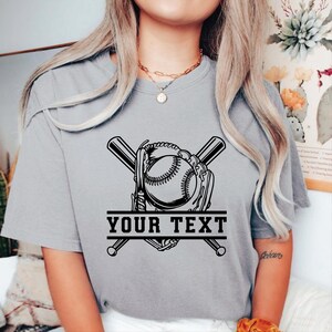 May include: A grey t-shirt with a baseball design. The graphic features a baseball, glove, and crossed bats, with the text "YOUR TEXT" in a banner below. The design is in black.