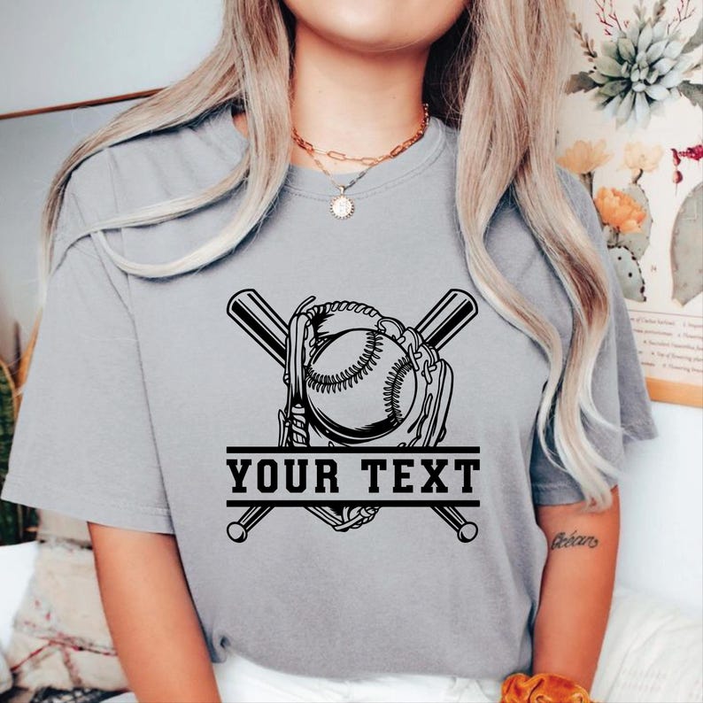 May include: A grey t-shirt with a baseball design. The graphic features a baseball, glove, and crossed bats, with the text "YOUR TEXT" in a banner below. The design is in black.