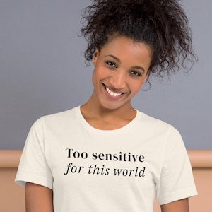 May include: Cream-colored t-shirt with the text "Too sensitive for this world" in black. The shirt has short sleeves and a crew neck. The model is wearing blue jeans.