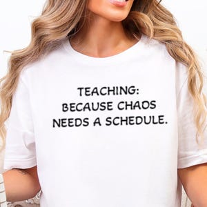 May include: A white short-sleeved t-shirt featuring black text centered on the chest that reads: TEACHING: BECAUSE CHAOS NEEDS A SCHEDULE. The shirt is worn with light blue denim jeans, and a white mug with a brown grid pattern is held in one hand.