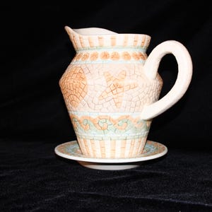 May include: A decorative ceramic pitcher with a matching saucer. The pitcher has a white handle and spout, and is decorated with a mosaic-like pattern in shades of peach, beige, and teal. The pitcher sits on a small, round saucer.