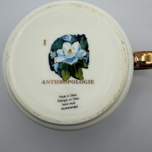 Puede incluir: Taza de cer&aacute;mica blanca con asa dorada. La base de la taza presenta un dise&ntilde;o floral con una flor azul y blanca y la palabra "ANTHROPOLOGIE". Tambi&eacute;n es visible el texto "Made in China, Fabriqu&eacute; en Chine, hand wash recommended".