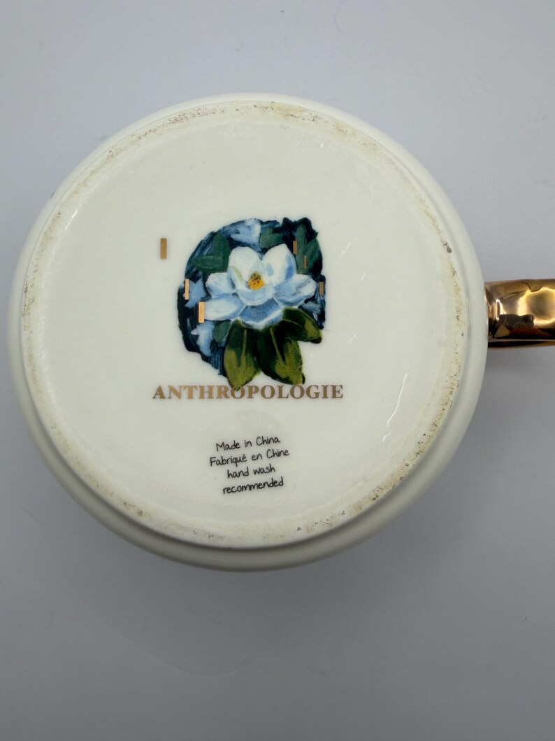 Puede incluir: Taza de cer&aacute;mica blanca con asa dorada. La base de la taza presenta un dise&ntilde;o floral con una flor azul y blanca y la palabra "ANTHROPOLOGIE". Tambi&eacute;n es visible el texto "Made in China, Fabriqu&eacute; en Chine, hand wash recommended".