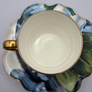 Puede incluir: Una taza blanca con asa y borde dorados se asienta sobre un platillo a juego. El platillo y la taza presentan un dise&ntilde;o floral abstracto azul y verde con ribete dorado. El juego est&aacute; dise&ntilde;ado para t&eacute; o caf&eacute;.