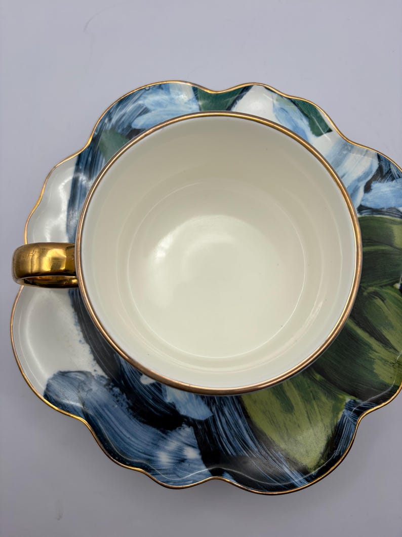 Puede incluir: Una taza blanca con asa y borde dorados se asienta sobre un platillo a juego. El platillo y la taza presentan un dise&ntilde;o floral abstracto azul y verde con ribete dorado. El juego est&aacute; dise&ntilde;ado para t&eacute; o caf&eacute;.