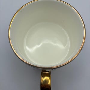 Puede incluir: Vista superior de una taza de t&eacute; blanca con borde y asa dorados. El interior de la taza es blanco y el asa es dorada. El borde de la taza tiene una fina banda dorada.
