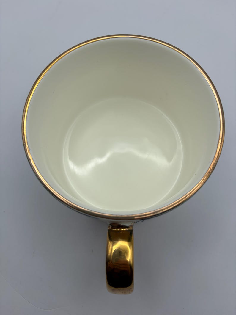 Puede incluir: Vista superior de una taza de t&eacute; blanca con borde y asa dorados. El interior de la taza es blanco y el asa es dorada. El borde de la taza tiene una fina banda dorada.