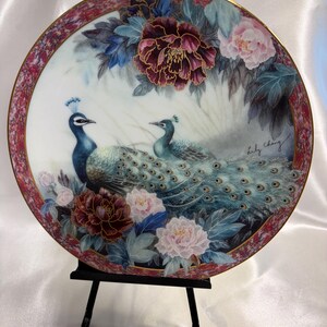May include: Decorative plate featuring two peacocks amidst floral arrangements. The plate has a maroon and gold border, with a central design of blue and teal peacocks, surrounded by burgundy and pink flowers. The plate is displayed on a black stand.
