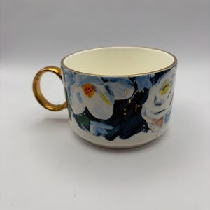 Puede incluir: Una taza de cer&aacute;mica con asa y borde dorados. La taza presenta un dise&ntilde;o floral con flores blancas, hojas azules y detalles dorados. El interior es de color crema y la taza est&aacute; sobre un fondo blanco.