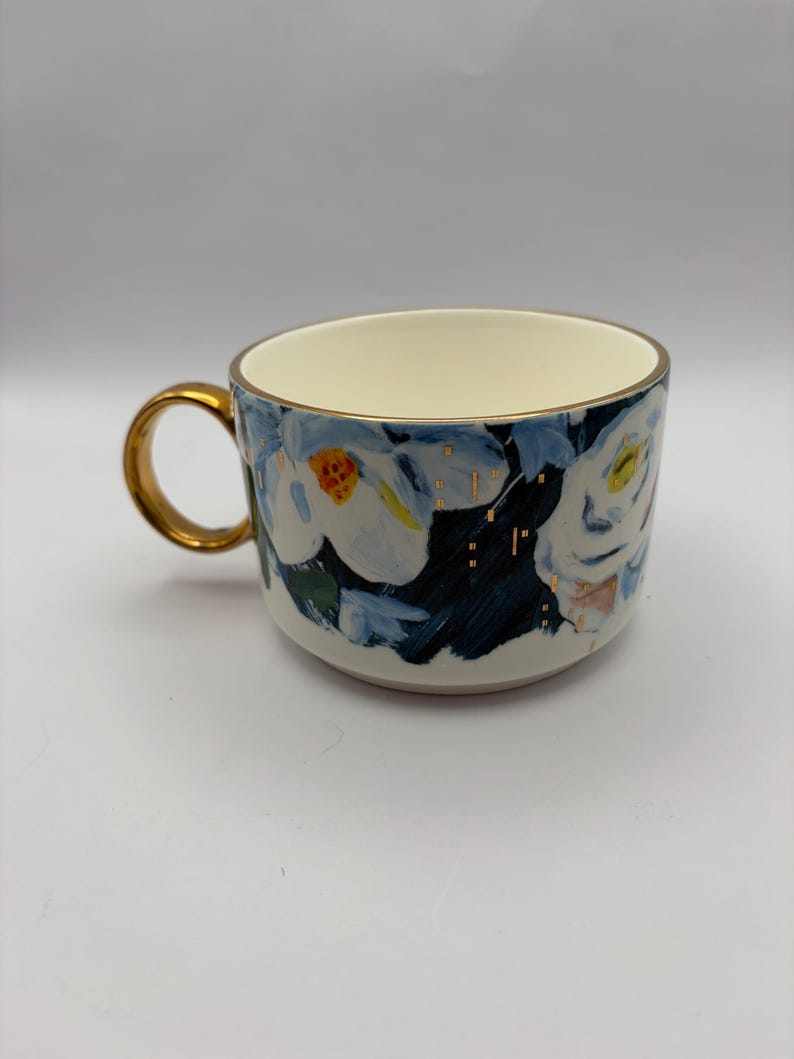 Puede incluir: Una taza de cer&aacute;mica con asa y borde dorados. La taza presenta un dise&ntilde;o floral con flores blancas, hojas azules y detalles dorados. El interior es de color crema y la taza est&aacute; sobre un fondo blanco.