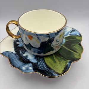 Puede incluir: Un juego de taza y platillo con motivos florales. La taza y el platillo presentan un dise&ntilde;o floral azul, blanco y verde con ribete dorado. La taza tiene un asa dorada y el platillo tiene un borde festoneado. El juego est&aacute; dise&ntilde;ado para t&eacute; o caf&eacute;.