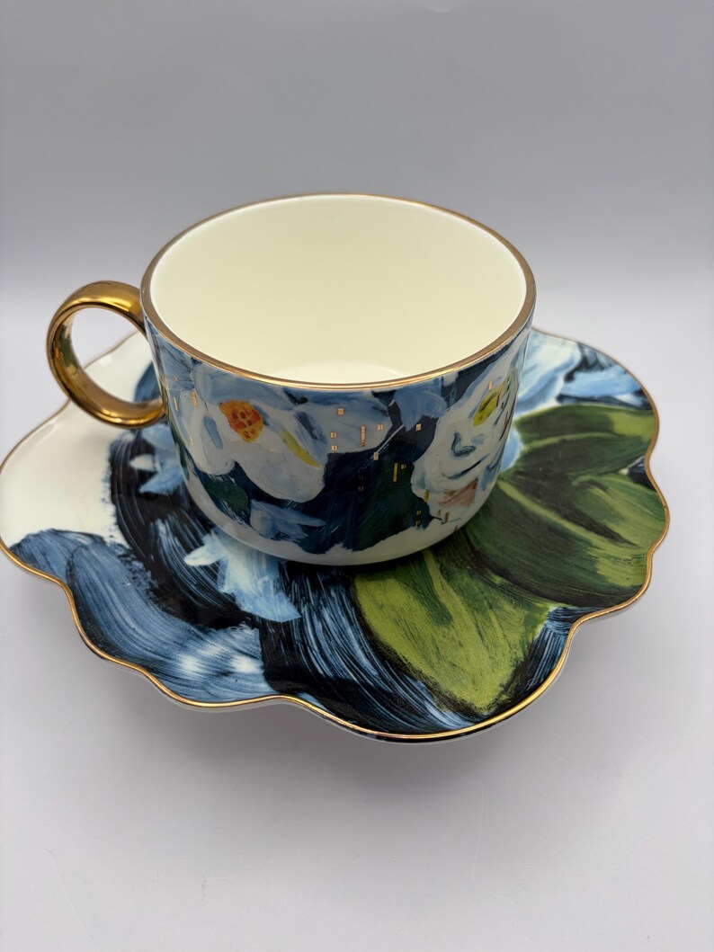 Puede incluir: Un juego de taza y platillo con motivos florales. La taza y el platillo presentan un dise&ntilde;o floral azul, blanco y verde con ribete dorado. La taza tiene un asa dorada y el platillo tiene un borde festoneado. El juego est&aacute; dise&ntilde;ado para t&eacute; o caf&eacute;.