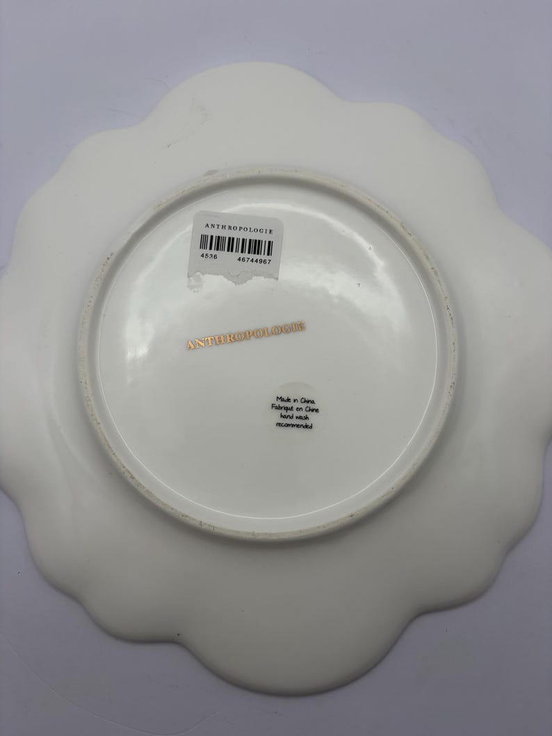 Puede incluir: Plato blanco de Anthropologie con borde festoneado. La parte inferior del plato presenta el nombre de la marca, una etiqueta con c&oacute;digo de barras y texto que indica que fue fabricado en China. El plato tiene un acabado liso y brillante.
