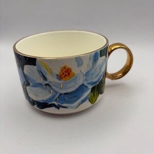 Puede incluir: Una taza de cer&aacute;mica con asa y borde dorados. La taza presenta un dise&ntilde;o pintado a mano de una flor azul y blanca con un centro amarillo. Acentos dorados est&aacute;n dispersos por la superficie de la taza, y el interior es de color crema.