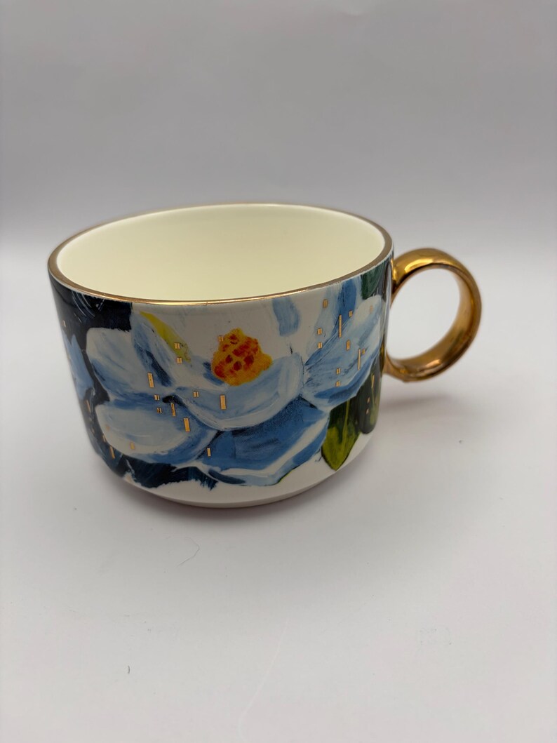 Puede incluir: Una taza de cer&aacute;mica con asa y borde dorados. La taza presenta un dise&ntilde;o pintado a mano de una flor azul y blanca con un centro amarillo. Acentos dorados est&aacute;n dispersos por la superficie de la taza, y el interior es de color crema.