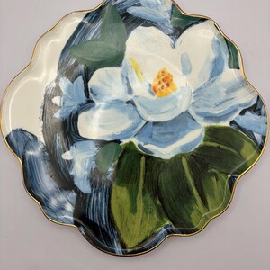 Puede incluir: Un plato decorativo con borde festoneado y borde dorado. El plato presenta un dise&ntilde;o pintado a mano de una gran flor de magnolia azul claro con hojas verdes sobre un fondo blanco y azul oscuro. Una pieza &uacute;nica de decoraci&oacute;n del hogar.