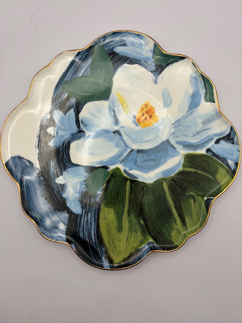 Puede incluir: Un plato decorativo con borde festoneado y borde dorado. El plato presenta un dise&ntilde;o pintado a mano de una gran flor de magnolia azul claro con hojas verdes sobre un fondo blanco y azul oscuro. Una pieza &uacute;nica de decoraci&oacute;n del hogar.