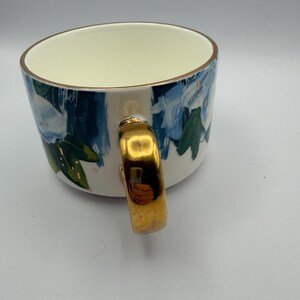 Puede incluir: Una taza de cer&aacute;mica blanca con asa y borde dorados. La taza est&aacute; decorada con motivos florales azules y hojas verdes. El interior de la taza es de color crema.