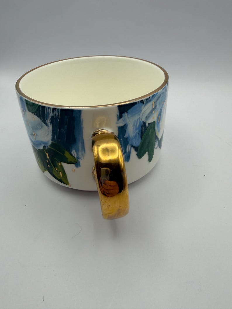Puede incluir: Una taza de cer&aacute;mica blanca con asa y borde dorados. La taza est&aacute; decorada con motivos florales azules y hojas verdes. El interior de la taza es de color crema.