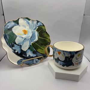 Puede incluir: Un juego de taza y platillo de cer&aacute;mica con un dise&ntilde;o floral. La taza y el platillo presentan un estampado floral blanco y azul con ribete dorado. El platillo tiene una forma &uacute;nica y festoneada. La taza tiene un asa dorada.