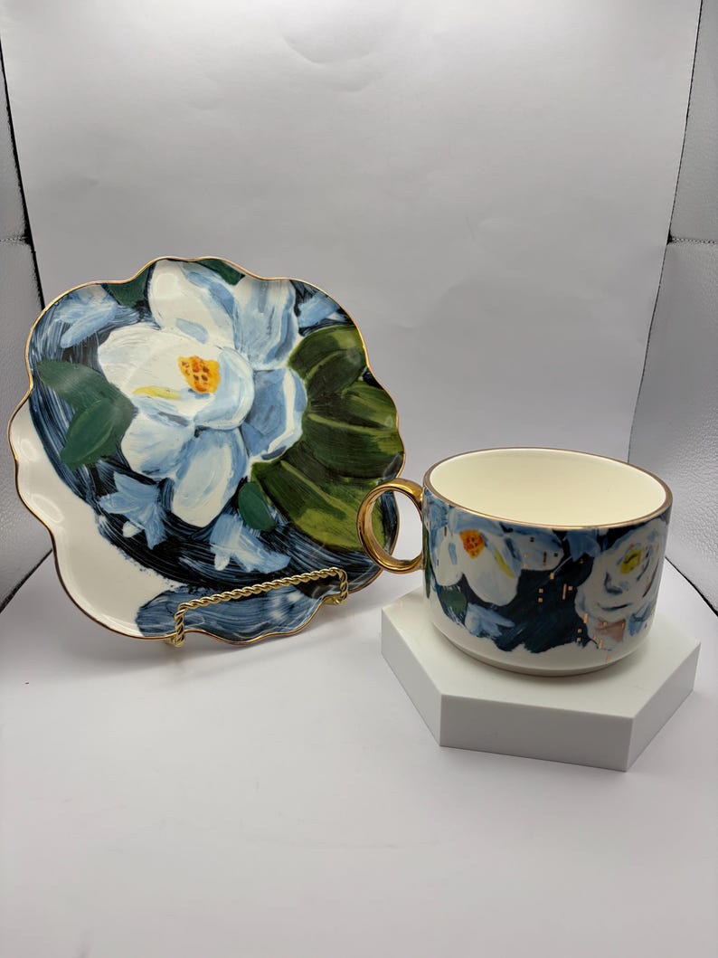 Puede incluir: Un juego de taza y platillo de cer&aacute;mica con un dise&ntilde;o floral. La taza y el platillo presentan un estampado floral blanco y azul con ribete dorado. El platillo tiene una forma &uacute;nica y festoneada. La taza tiene un asa dorada.