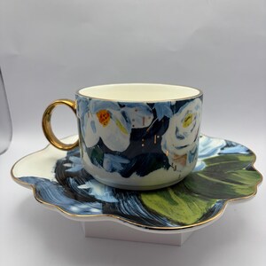 Puede incluir: Un juego de taza y platillo de cer&aacute;mica. La taza y el platillo presentan un dise&ntilde;o floral con detalles en azul, blanco y dorado. La taza tiene un asa y un borde dorados. El platillo tiene forma de flor con hojas pintadas de verde y azul.