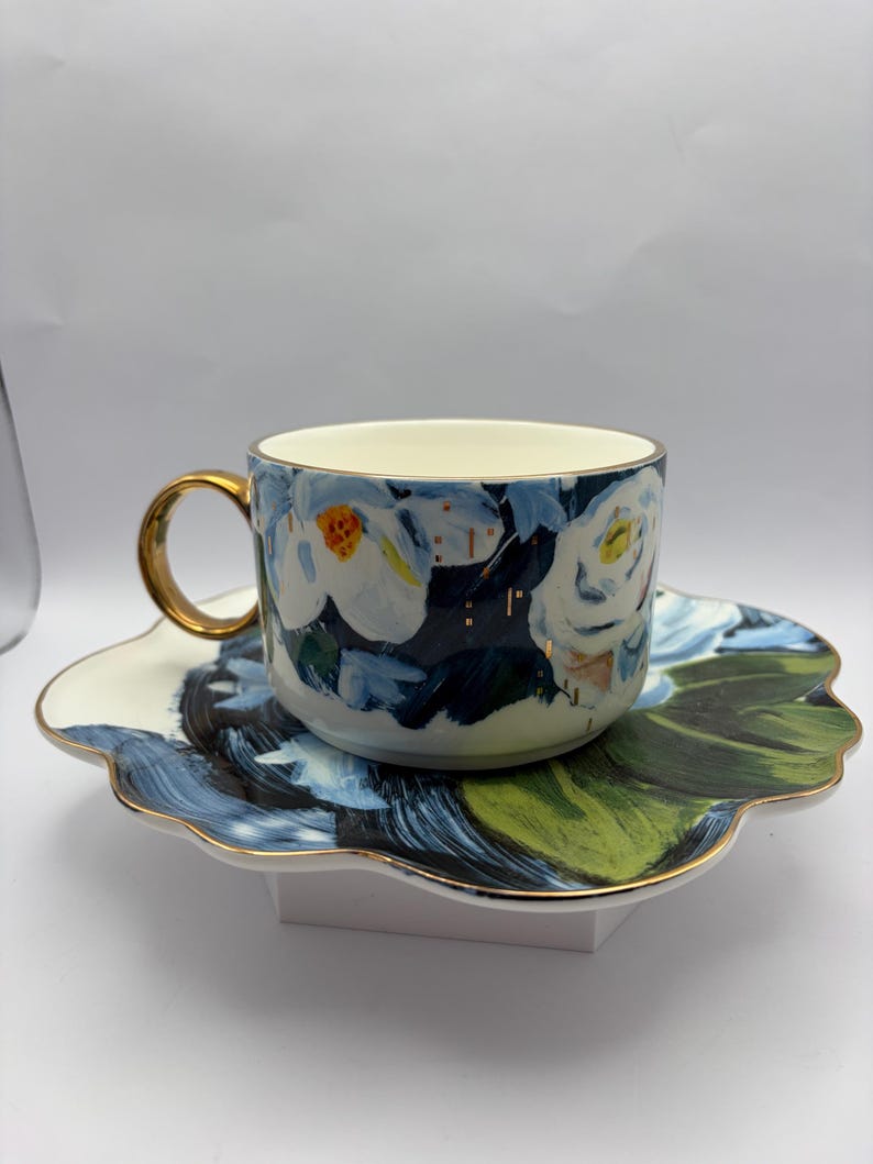 Puede incluir: Un juego de taza y platillo de cer&aacute;mica. La taza y el platillo presentan un dise&ntilde;o floral con detalles en azul, blanco y dorado. La taza tiene un asa y un borde dorados. El platillo tiene forma de flor con hojas pintadas de verde y azul.