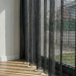 Könnte beinhalten: Ein bodenlanger schwarzer Vorhang mit einem gitterartigen, offen gewebten Netzmuster. Der Stoff fällt in vertikalen Falten und lässt Sonnenlicht durch die Lücken scheinen, wodurch ein markantes, quadratisches Schattenmuster auf dem Holzboden entsteht.