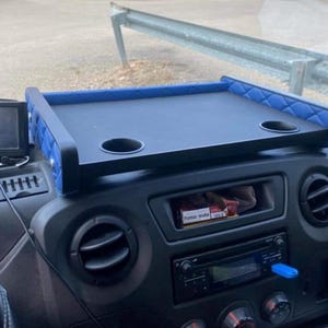 Peut inclure: Plateau de tableau de bord noir avec des bords matelassés bleus et deux porte-gobelets. Un appareil GPS Garmin est monté sur le tableau de bord. Le plateau est situé au-dessus de la radio et des bouches d'aération. Une clé USB bleue est branchée sur la radio.