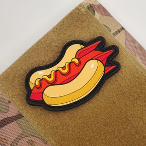 May include: A colorful hot dog patch with a black border. The hot dog features a yellow bun, a red hot dog, and yellow mustard. The patch is on a tan and camouflage background.