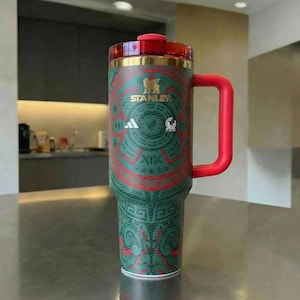 Puede incluir: Un vaso t&eacute;rmico verde y rojo con asa y tapa rojas. El vaso presenta un dise&ntilde;o decorativo de inspiraci&oacute;n azteca, el logotipo de Stanley y el logotipo de Adidas. El vaso est&aacute; dise&ntilde;ado para mantener las bebidas fr&iacute;as o calientes.