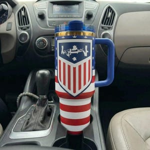 May include: A tall, cylindrical insulated tumbler with a blue handle and a lid. The exterior features a patriotic design with red and white horizontal stripes on the bottom and a blue shield graphic with white stars and stripes on the upper portion.