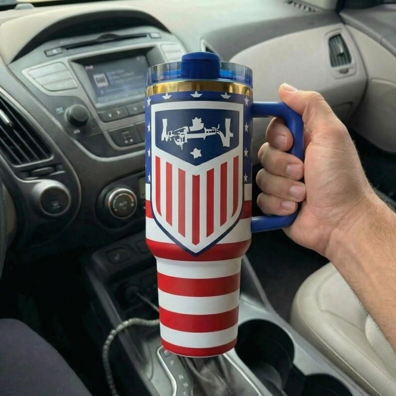 May include: A tall insulated tumbler with a blue handle and a clear lid, featuring a patriotic design with red and white horizontal stripes on the bottom and a blue shield graphic with white stars and a central white rifle silhouette on top.