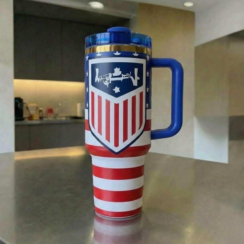 May include: A tall, insulated stainless steel tumbler with a blue handle and lid. The exterior features a red, white, and blue American flag-inspired design with a central shield emblem containing a white graphic of a rifle and a star.