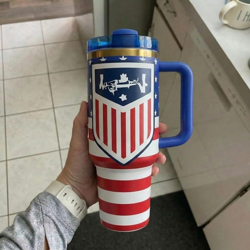 May include: A large insulated tumbler with a handle, featuring a blue, white, and red American flag pattern. The front displays a shield-shaped emblem with a white graphic of a rifle and stars. It has a blue lid and a gold-colored rim band.