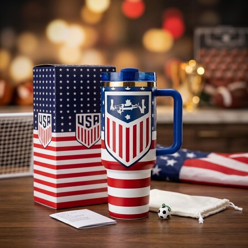 May include: A tall insulated tumbler with a handle, featuring a red, white, and blue USA soccer crest and flag pattern. It includes a matching box with the same design, a small soccer ball charm, and a white drawstring pouch on a wooden surface.