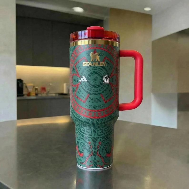 Puede incluir: Un vaso t&eacute;rmico verde y rojo con asa y tapa rojas. El vaso presenta un dise&ntilde;o decorativo de inspiraci&oacute;n azteca, el logotipo de Stanley y el logotipo de Adidas. El vaso est&aacute; dise&ntilde;ado para mantener las bebidas fr&iacute;as o calientes.