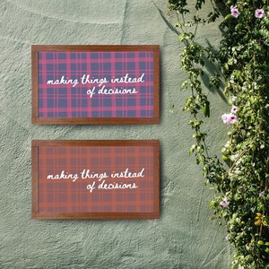 May include: Two framed art prints with plaid patterns and the text "making things instead of decisions." The top print has a blue and pink plaid, while the bottom print has an orange and red plaid. The frames are brown.