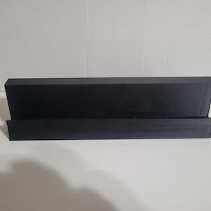 May include: A black, rectangular display stand with a sloped back and a lower ledge. The stand is made of a solid, matte material and is designed to hold items. The stand is on a white surface.