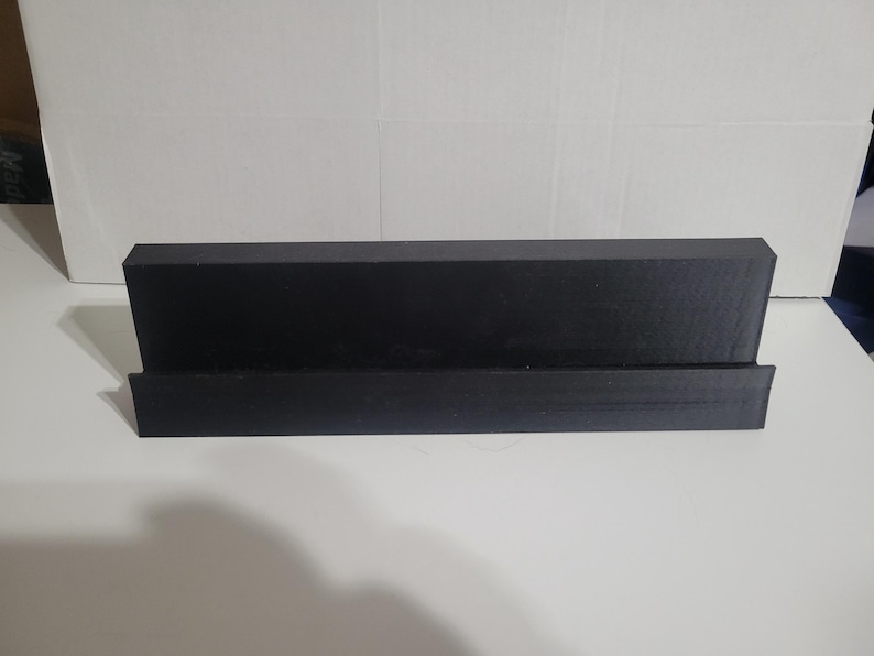 May include: A black, rectangular display stand with a sloped back and a lower ledge. The stand is made of a solid, matte material and is designed to hold items. The stand is on a white surface.
