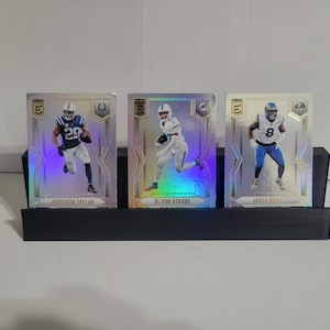 May include: A black, three-slot card display holding three football trading cards. The cards feature players in action, with team colors and player names visible. The cards have a holographic sheen.