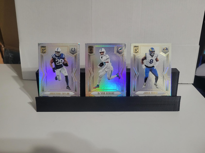 May include: A black, three-slot card display holding three football trading cards. The cards feature players in action, with team colors and player names visible. The cards have a holographic sheen.