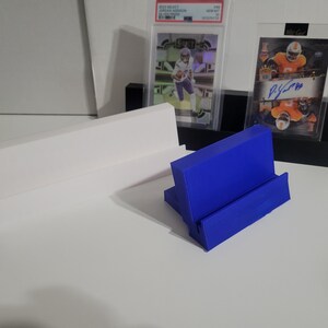 May include: A blue card display stand is in the foreground, with a white and black display stand behind it. Two trading cards are displayed in protective cases. The background is a white surface.