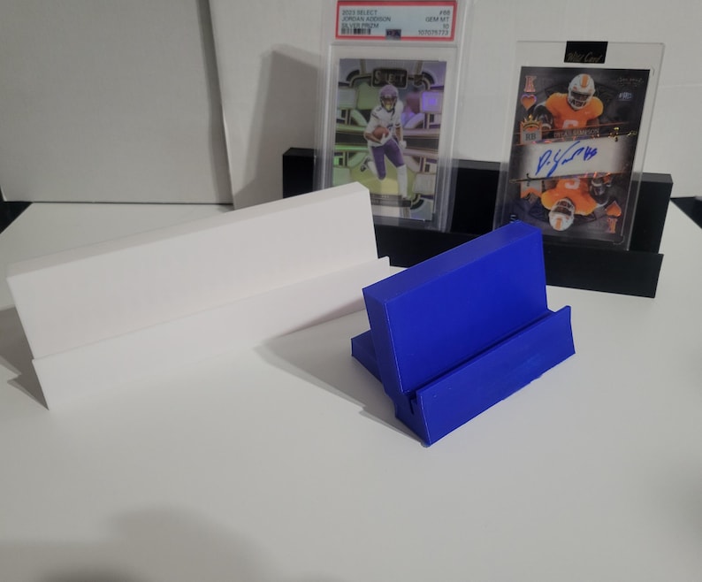 May include: A blue card display stand is in the foreground, with a white and black display stand behind it. Two trading cards are displayed in protective cases. The background is a white surface.