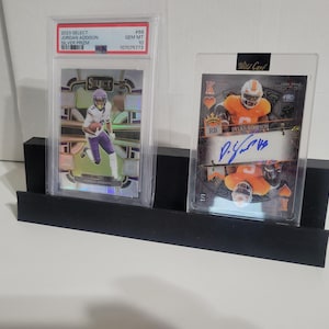 May include: A black display stand holding two sports trading cards in protective cases. One card features a football player in purple and white, the other an orange and white player with an autograph. The stand is rectangular.