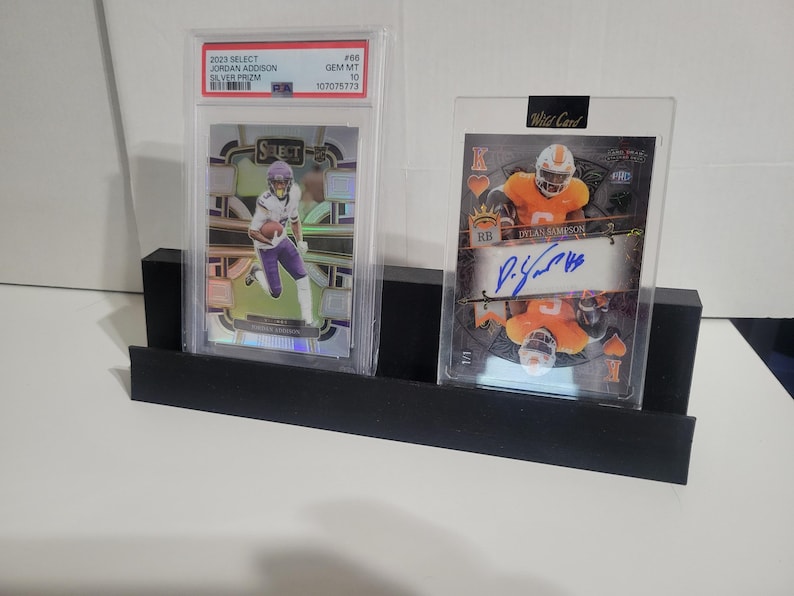May include: A black display stand holding two sports trading cards in protective cases. One card features a football player in purple and white, the other an orange and white player with an autograph. The stand is rectangular.