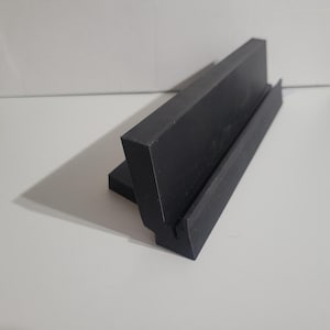 May include: A black, angled display stand with a groove, likely for holding a phone or small item. The stand is made of a solid, dark material and sits on a white surface. The design is simple and modern.