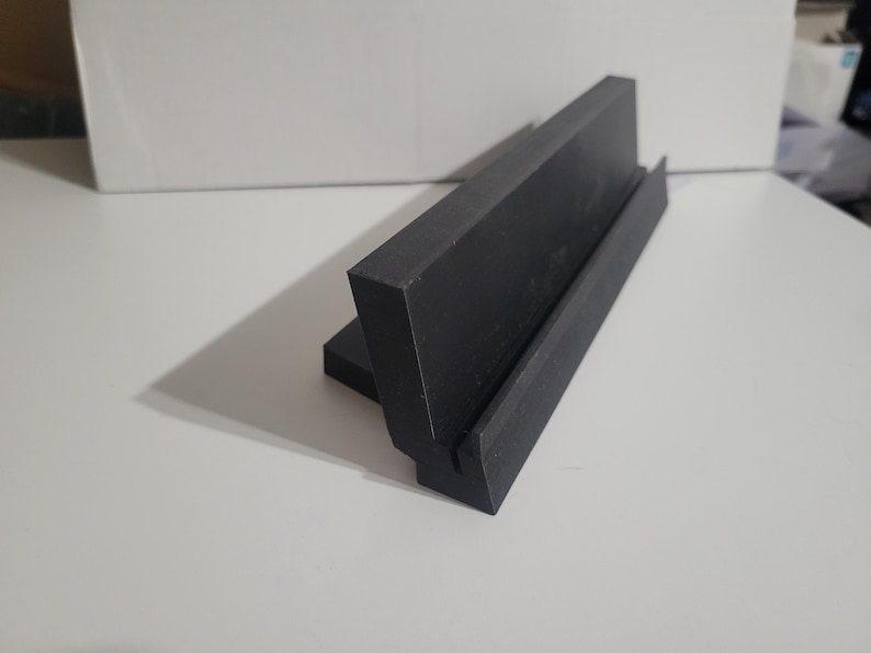 May include: A black, angled display stand with a groove, likely for holding a phone or small item. The stand is made of a solid, dark material and sits on a white surface. The design is simple and modern.